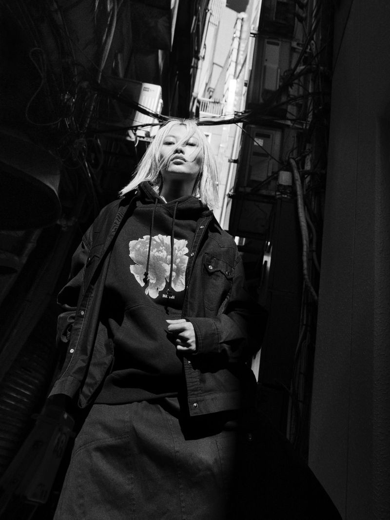 Black and white portrait of an adult female in a hooded sweatshirt and jacket standing in an alleyway with overhead wiring.