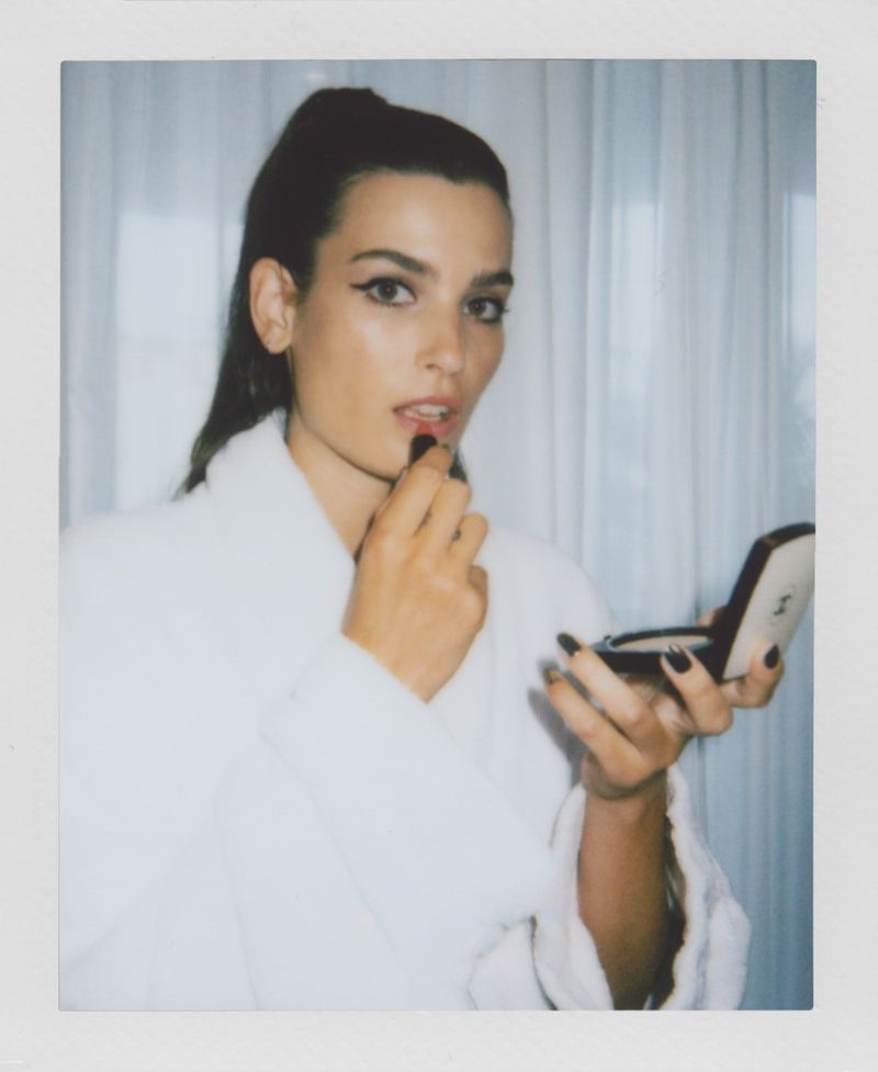 Portrait of a woman in a white robe applying red lipstick while holding a Chanel compact mirror indoors in a brightly lit room.
