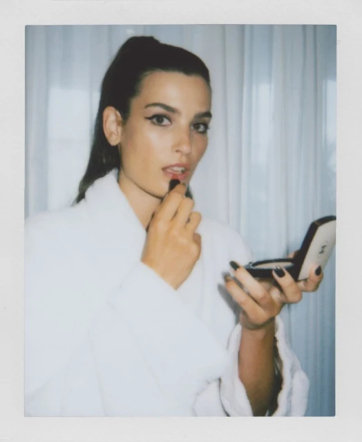 Portrait of a woman in a white robe applying red lipstick while holding a Chanel compact mirror indoors in a brightly lit room.