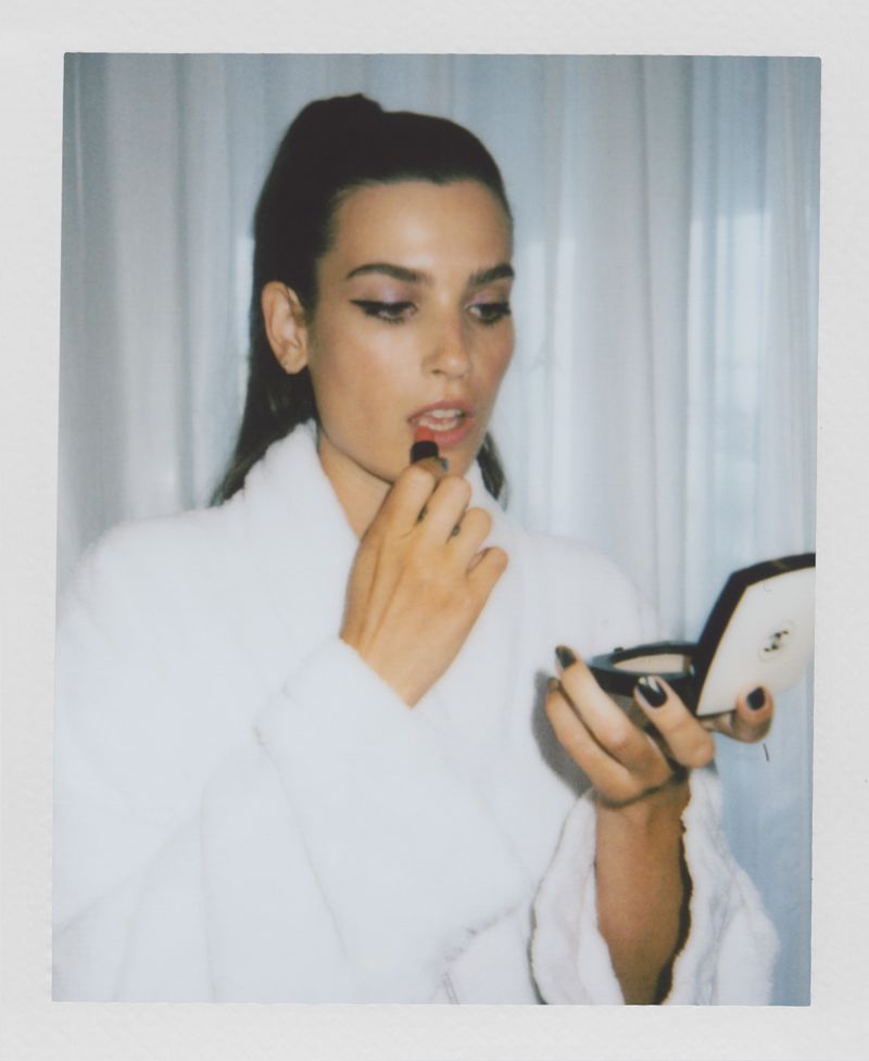 Brunette woman in white robe applies red lipstick, holding a compact mirror in an indoor setting with sheer white curtains.