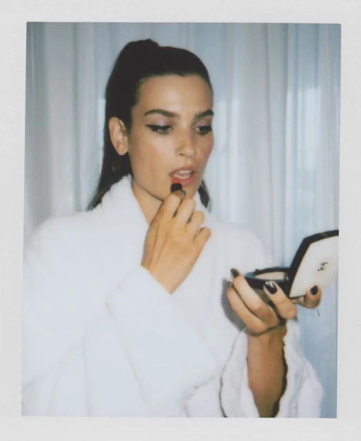 Brunette woman in white robe applies red lipstick, holding a compact mirror in an indoor setting with sheer white curtains.