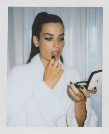 Brunette woman in white robe applies red lipstick, holding a compact mirror in an indoor setting with sheer white curtains.