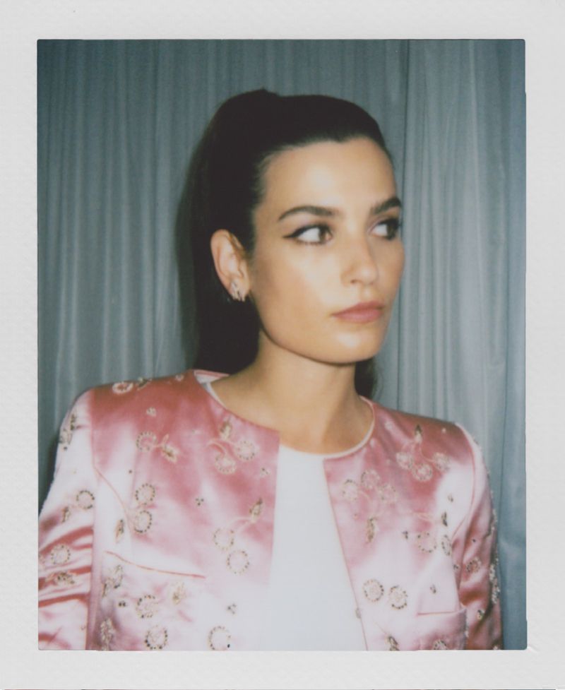 Portrait of a young woman with dark hair, wearing a pink floral jacket, looking to the side against a neutral backdrop.
