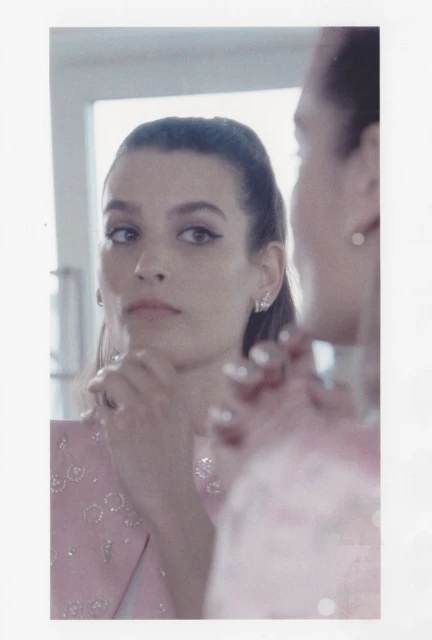 Close-up portrait of a stylish adult woman with dark hair looking into a mirror, wearing a light pink jacket indoors.