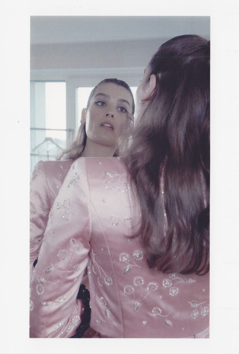 Woman gazes in the mirror, wearing a pink jacket with floral details. Interior shot with natural light, creating a soft, reflective ambiance.