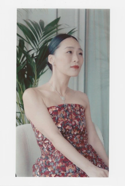 A fashionable Asian woman poses indoors, wearing a strapless floral dress and elegant jewelry, against a neutral background with green plants.