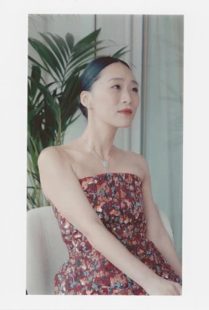 A fashionable Asian woman poses indoors, wearing a strapless floral dress and elegant jewelry, against a neutral background with green plants.