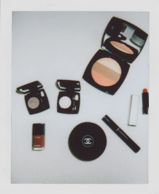 Composition of Chanel makeup products and nail polish in brown and black, arranged on a light surface in a polaroid-style frame.