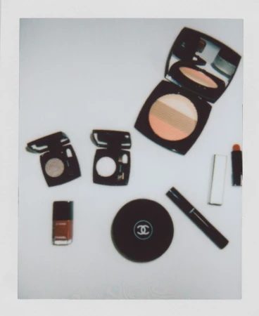Composition of Chanel makeup products and nail polish in brown and black, arranged on a light surface in a polaroid-style frame.