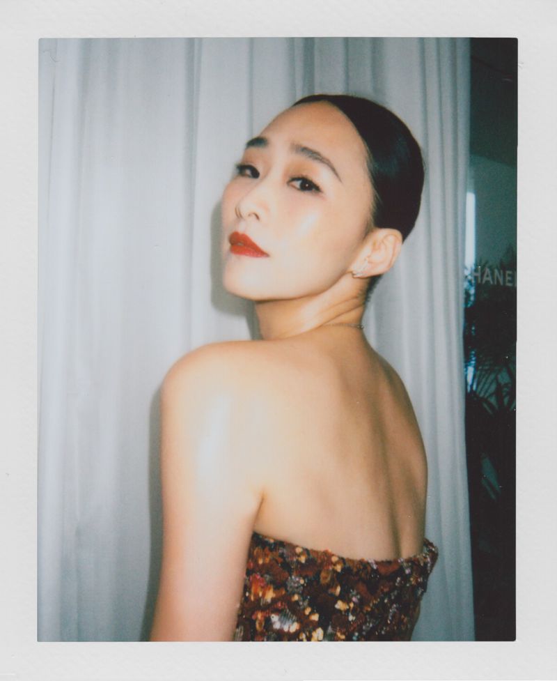 Portrait of an Asian woman with dark hair pulled back, wearing dark sequined top, looking over her shoulder toward the viewer in indoor setting.