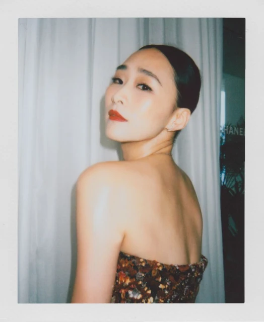 Portrait of an Asian woman with dark hair pulled back, wearing dark sequined top, looking over her shoulder toward the viewer in indoor setting.