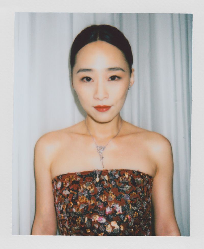 Elegant portrait of an Asian woman with dark hair, floral top, and a necklace, posing against a white curtain in soft lighting.