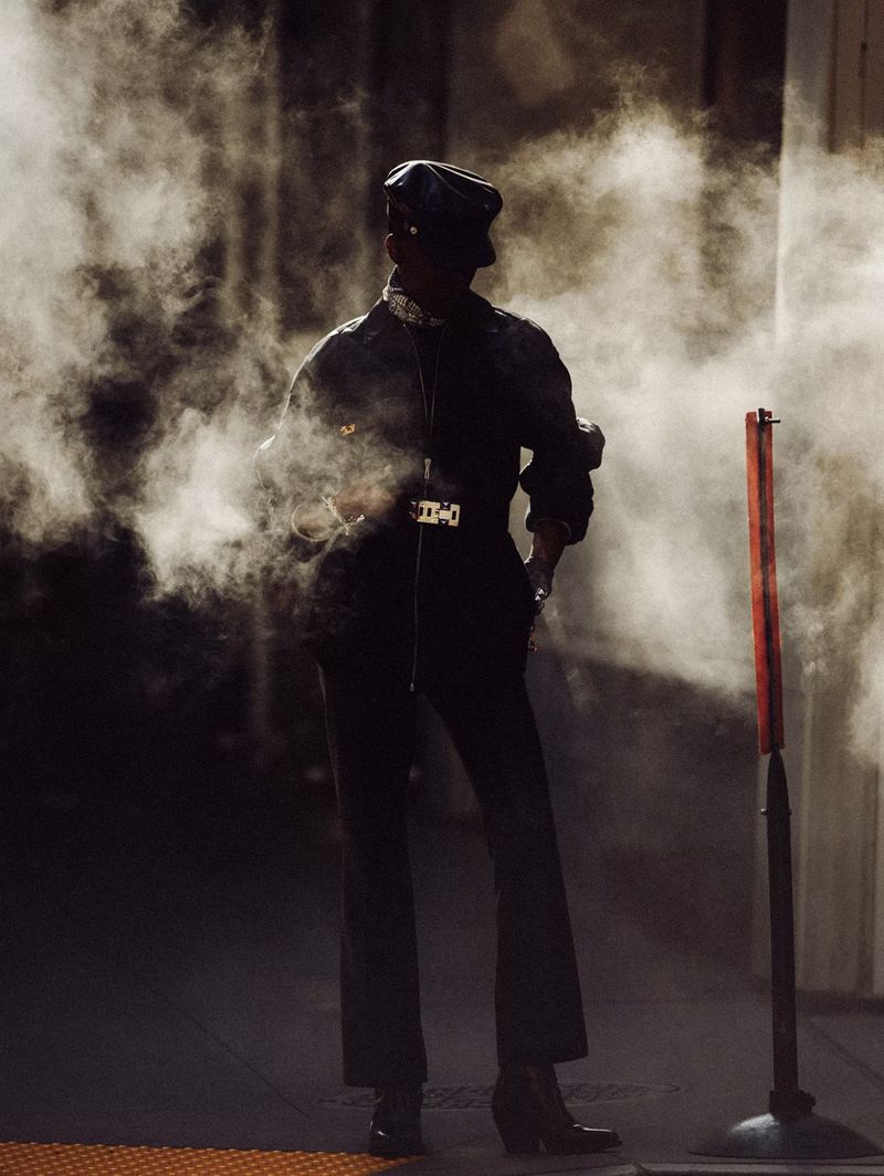 A full-length shot of an adult standing outdoors in a black jumpsuit and hat, partially obscured by mist, with a vintage fashion editorial look.