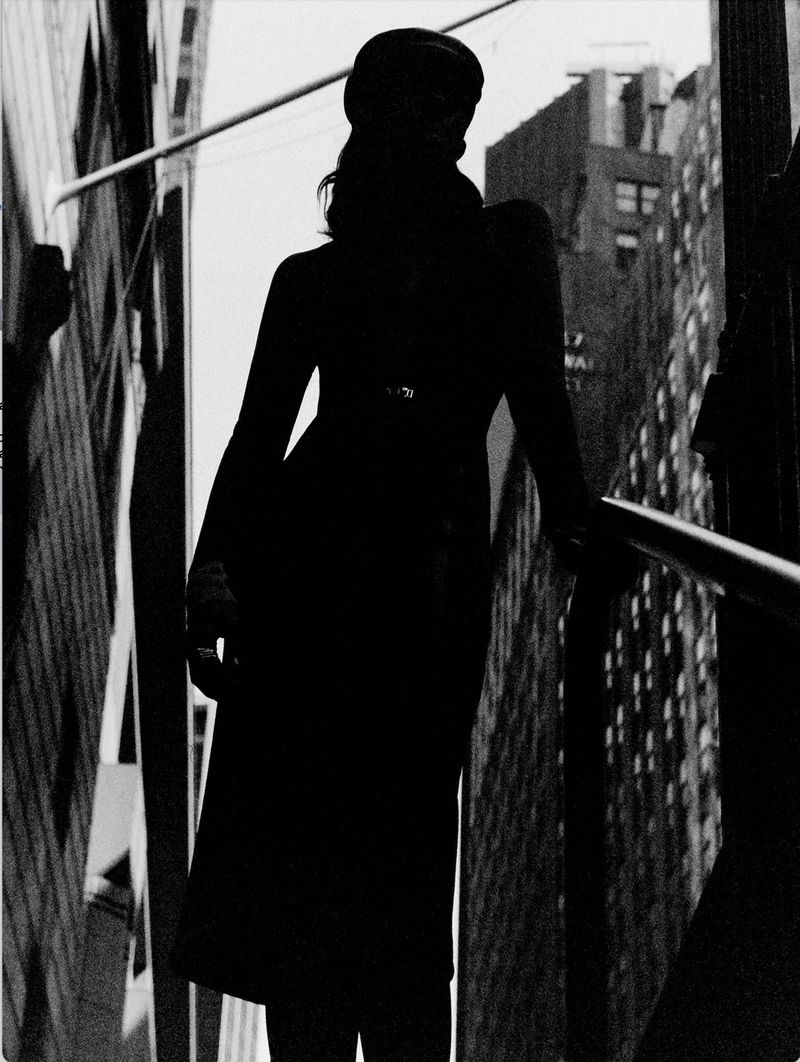 Monochrome silhouette of an adult woman in a dress and hat, posing outdoors with high-rise buildings in the blurred background.