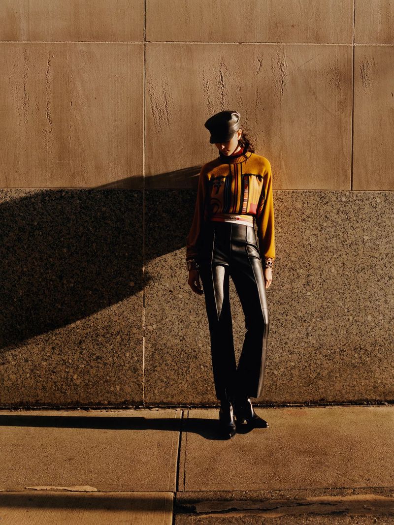A woman poses in an urban setting wearing a yellow patterned top, black leather pants, a black hat, and black boots against a wall.