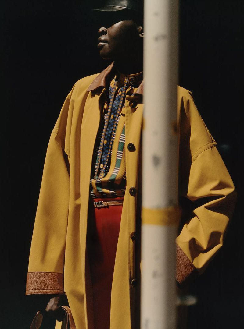 Woman in a yellow coat and leather cap standing near a pole looking off into the distance in a high fashion outdoor setting.