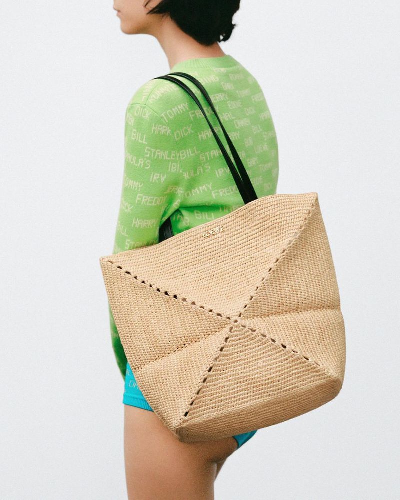 Woman showcases a large woven tote bag with geometric patchwork design, wearing a light green sweater and blue shorts against a neutral backdrop.