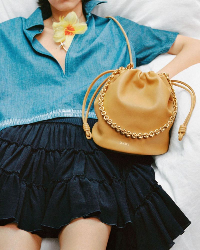 Fashion layout featuring a woman in a denim top and ruffled skirt with a tan Loewe handbag, lying on a white surface with a daffodil.