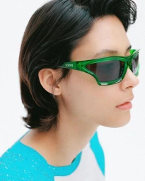 Close-up portrait of a model wearing translucent green sunglasses and a turquoise raglan shirt against a white backdrop.
