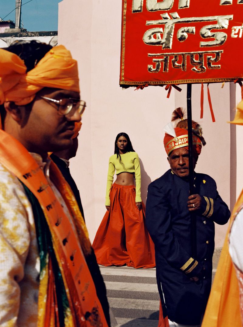 Woman in orange skirt and lime top standing in front of Indian band members with festive turbans and signage in an outdoor setting.