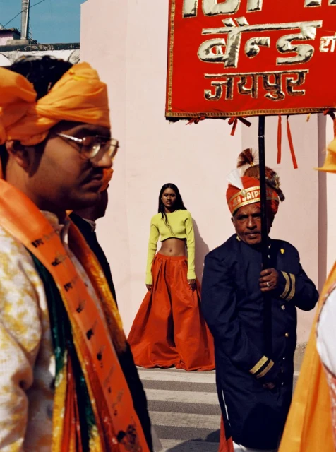 Woman in orange skirt and lime top standing in front of Indian band members with festive turbans and signage in an outdoor setting.