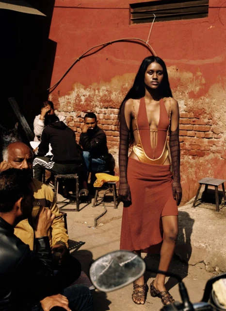 A striking fashion photograph featuring an adult female model in a terracotta dress, posing against an aged brick wall in natural light.