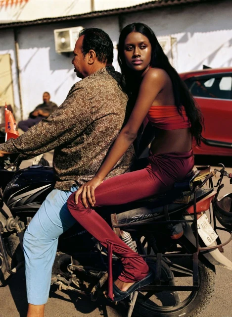 Woman poses on a motorbike while a driver navigates a city street, showcasing a blend of fashion and urban transportation.