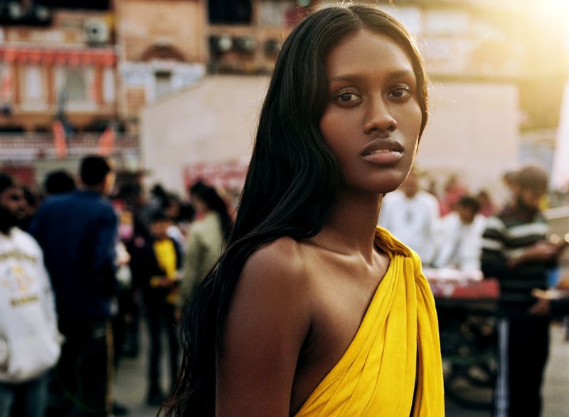 Close-up of a dark-skinned woman wearing a yellow dress, with dark hair, set against a blurred urban background with people and buildings.
