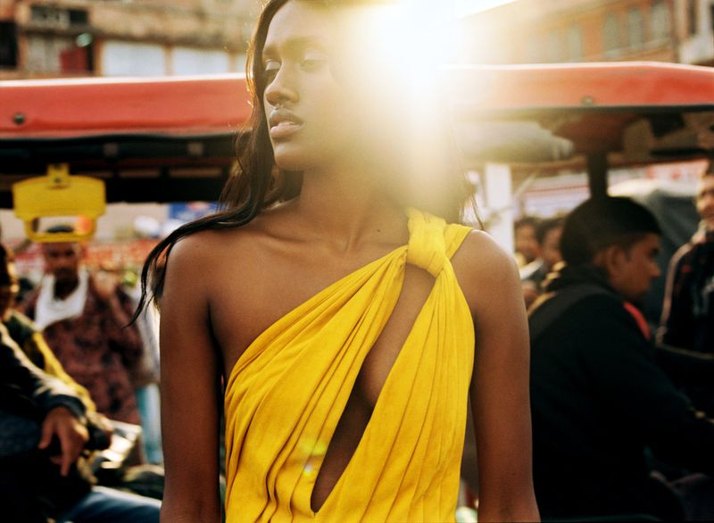 Elegant woman in a yellow dress stands outdoors in an urban setting with other people visible in the background and bright sunlight.