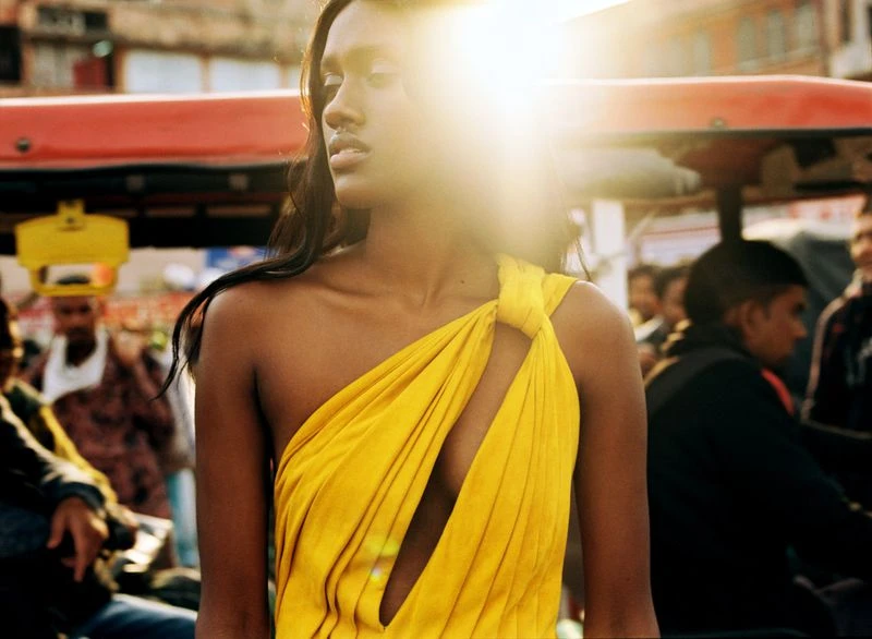 Elegant woman in a yellow dress stands outdoors in an urban setting with other people visible in the background and bright sunlight.