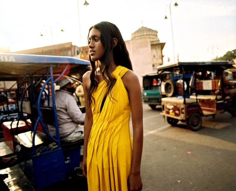 A woman in a yellow dress stands in a bustling city street with motorized rickshaws, under clear skies and warm lighting.