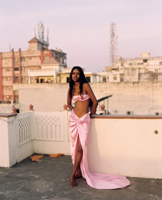 Woman in pink two-piece outfit poses on rooftop with buildings in the background, bathed in soft, warm light during daytime.