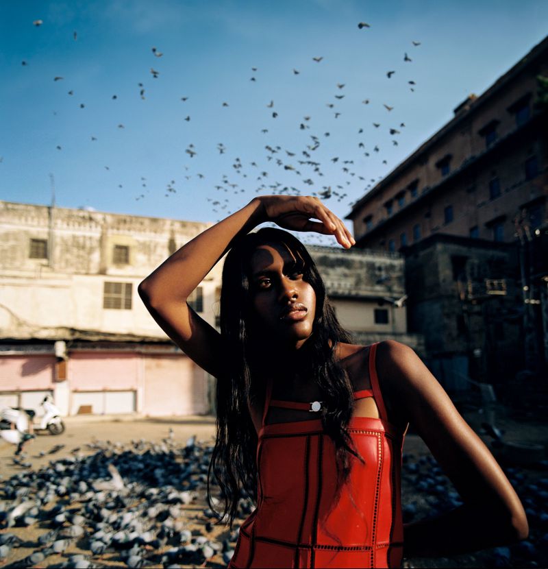 Fashion shot of a dark-skinned woman in a red dress, posing in a plaza surrounded by flocks of birds and old buildings under a bright sky.