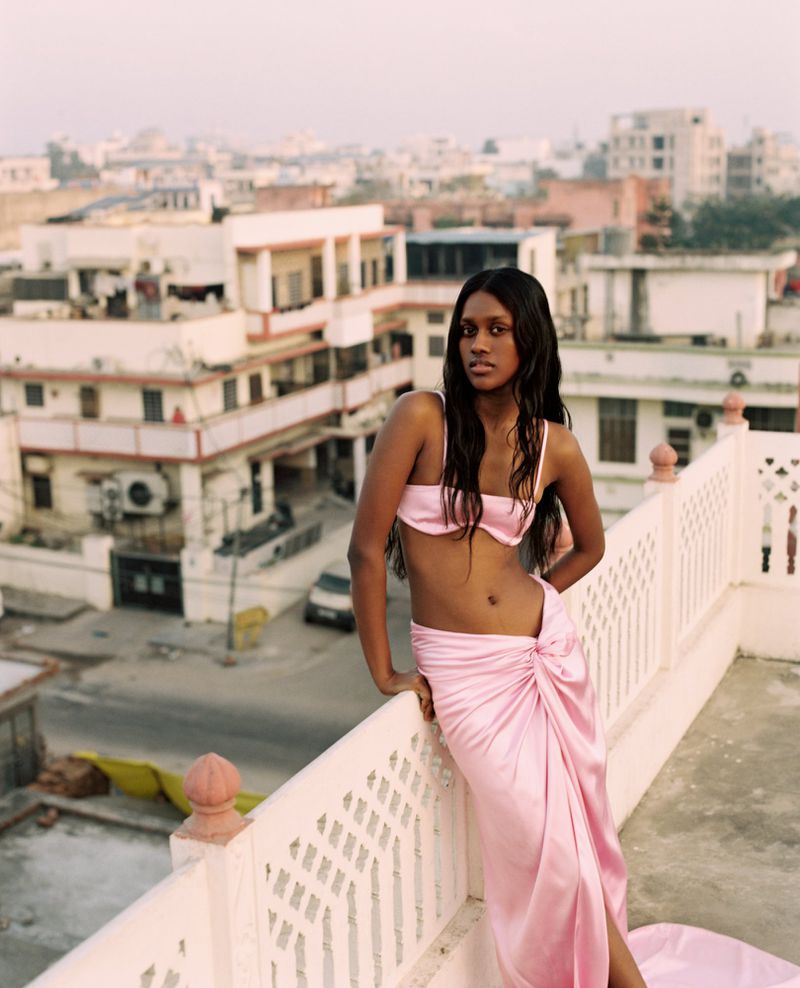 A striking portrait of a woman wearing a pink skirt and top, posing against a cityscape on a rooftop terrace with warm lighting.