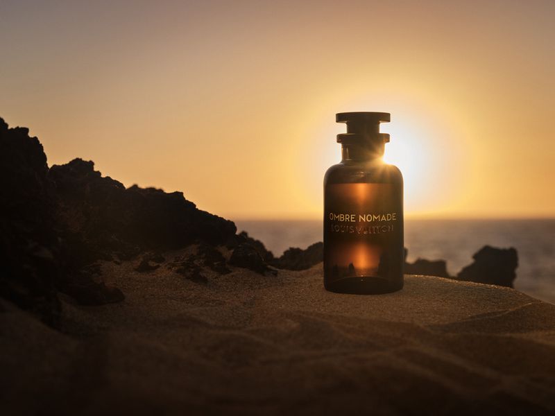 Louis Vuitton Ombre Nomade perfume bottle on a sandy surface at sunset, with the ocean visible in the background and golden light.