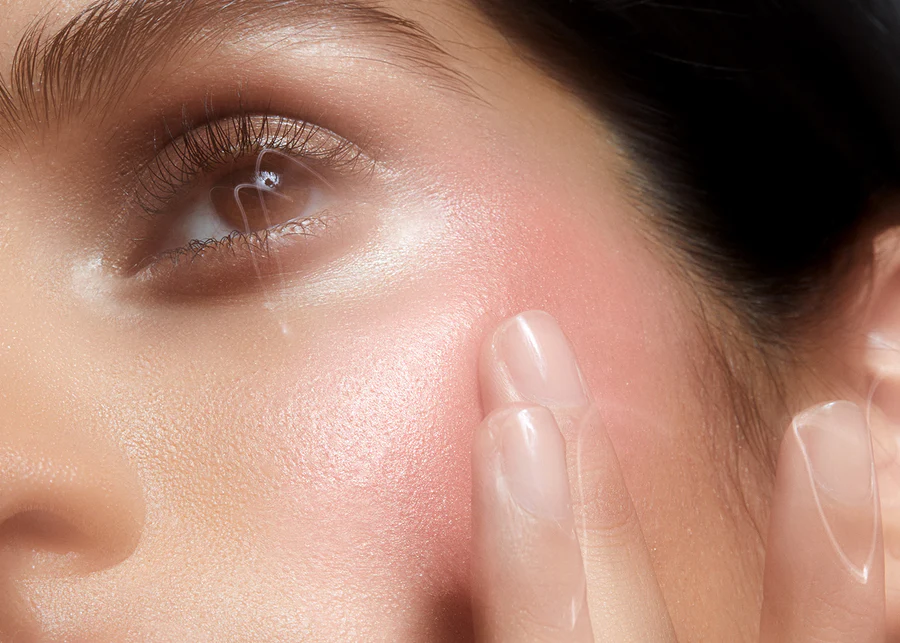 Close-up of a woman's face with pink blush. Her eye is open, and fingers are visible at her cheek in a beauty context.