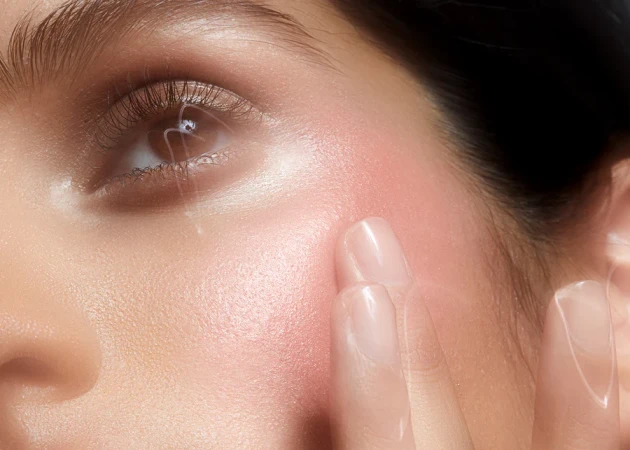 Close-up of a woman's face with pink blush. Her eye is open, and fingers are visible at her cheek in a beauty context.