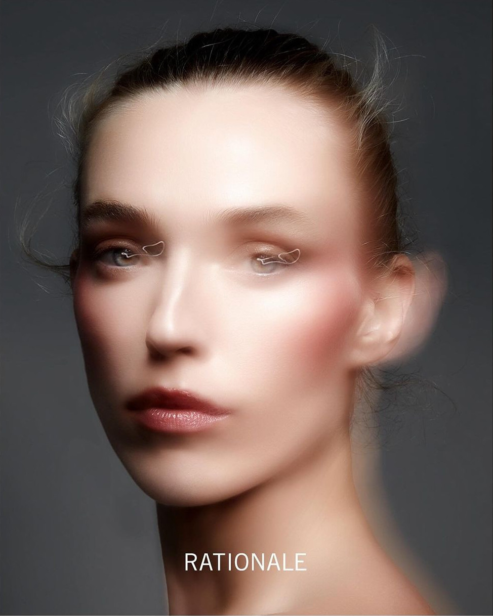 Close-up portrait of an adult female with light skin and blurred features, wearing pink blush, against a gray studio backdrop.