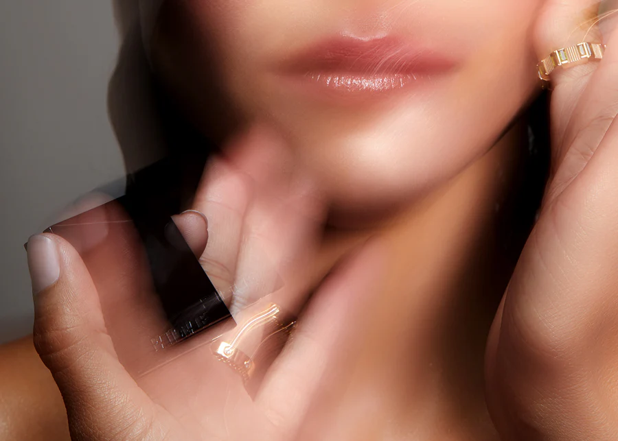 Close-up of an adult female's face with soft lighting, subtle makeup, a blurred hand, and gold jewelry, creating a beauty-focused composition.