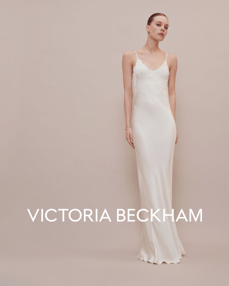 A woman in a sleeveless white gown stands in front of a beige background, in an indoor setting. The dress features delicate lace accents.