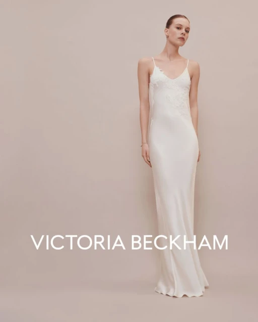 A woman in a sleeveless white gown stands in front of a beige background, in an indoor setting. The dress features delicate lace accents.