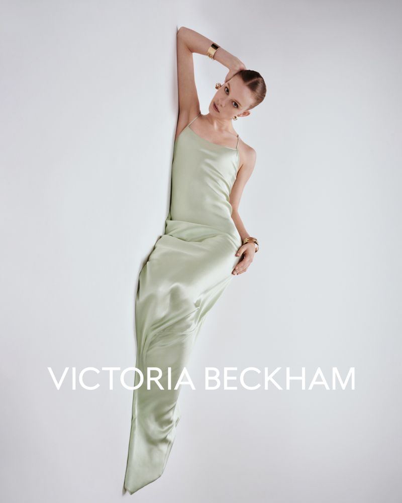 A slender fashion model leans against a white wall, wearing a long, flowing light green satin dress, gold earrings, and a gold bracelet.