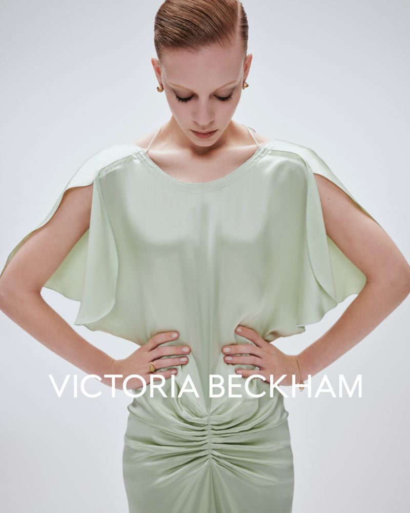 Fashion shot of a woman with hands on her waist wearing a light green dress with gathered fabric details, studio lighting and a white background.