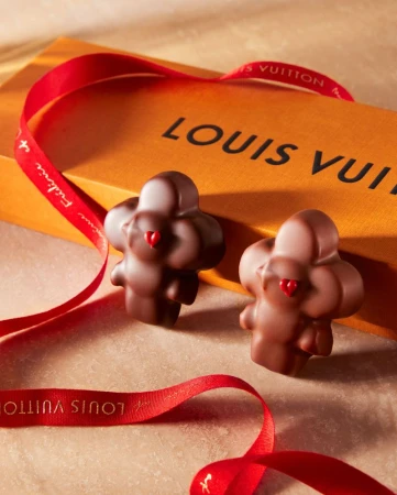 Two flower-shaped chocolates sit beside a Louis Vuitton box and red ribbon, evoking luxury confectionery and a sweet indulgence.