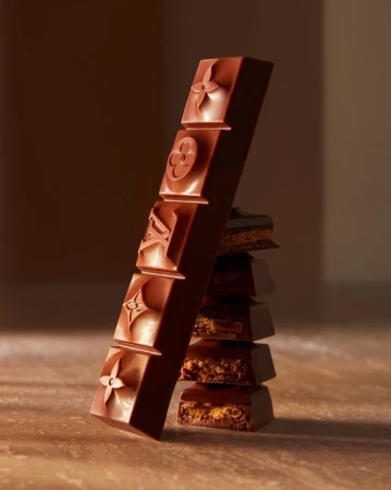 Close-up of a tall chocolate bar with geometric patterns leaning against a stacked pile of chocolate squares in warm studio light.