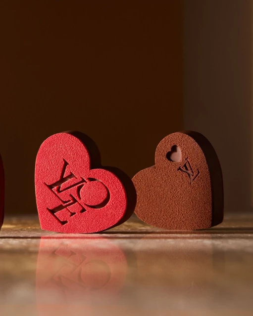 Three heart-shaped objects, one red with 'LOVE' carved, another brown with a logo, against a diffused brown background.