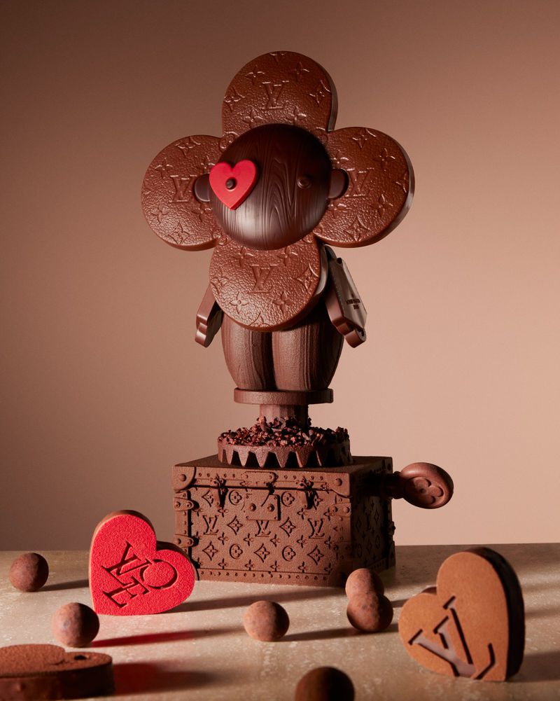 Chocolate sculpture of a flower-headed figure with Louis Vuitton branding and heart details, set against a neutral beige background.