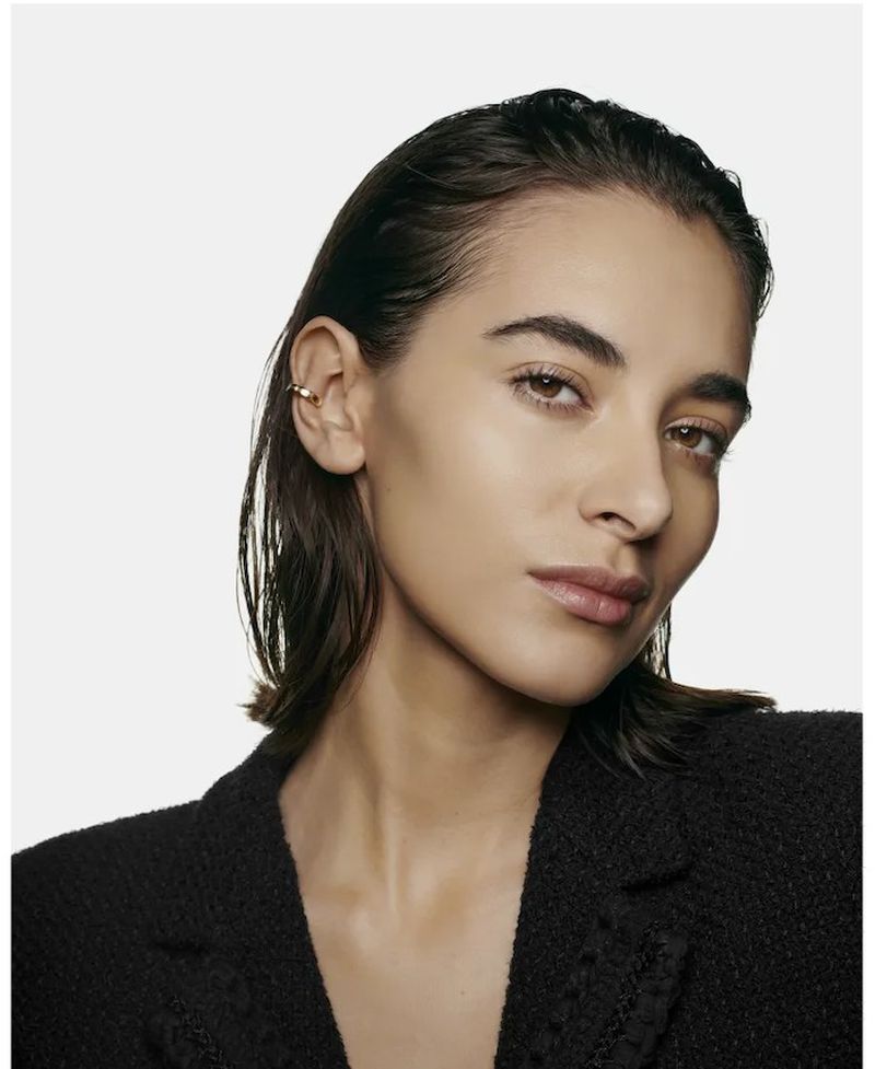 Close-up portrait of a young adult woman with slicked-back brunette hair, wearing a black jacket and gold ear cuff.