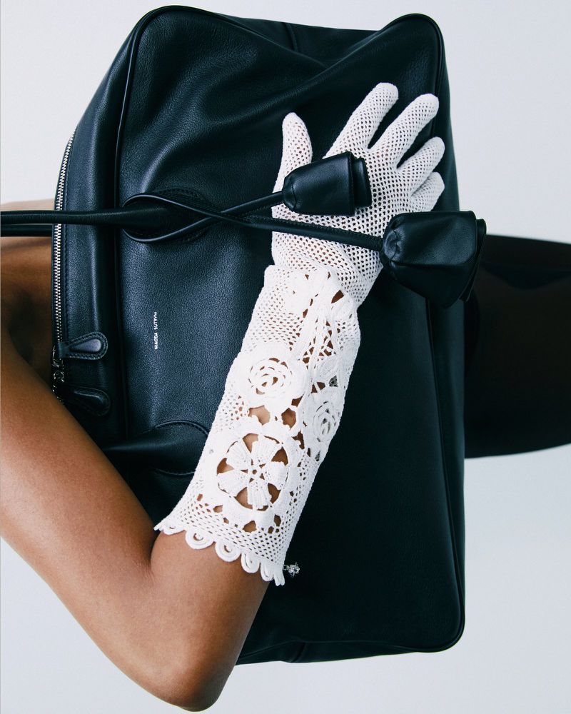 Close-up of a black leather handbag held by an arm wearing an ornate, crocheted white glove on a white backdrop.