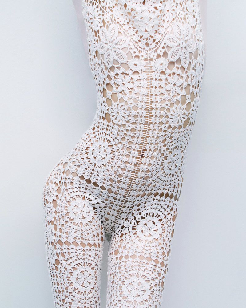 Close-up of a person wearing a white, intricate lace bodysuit against a light background, showcasing the garment's floral and geometric patterns.
